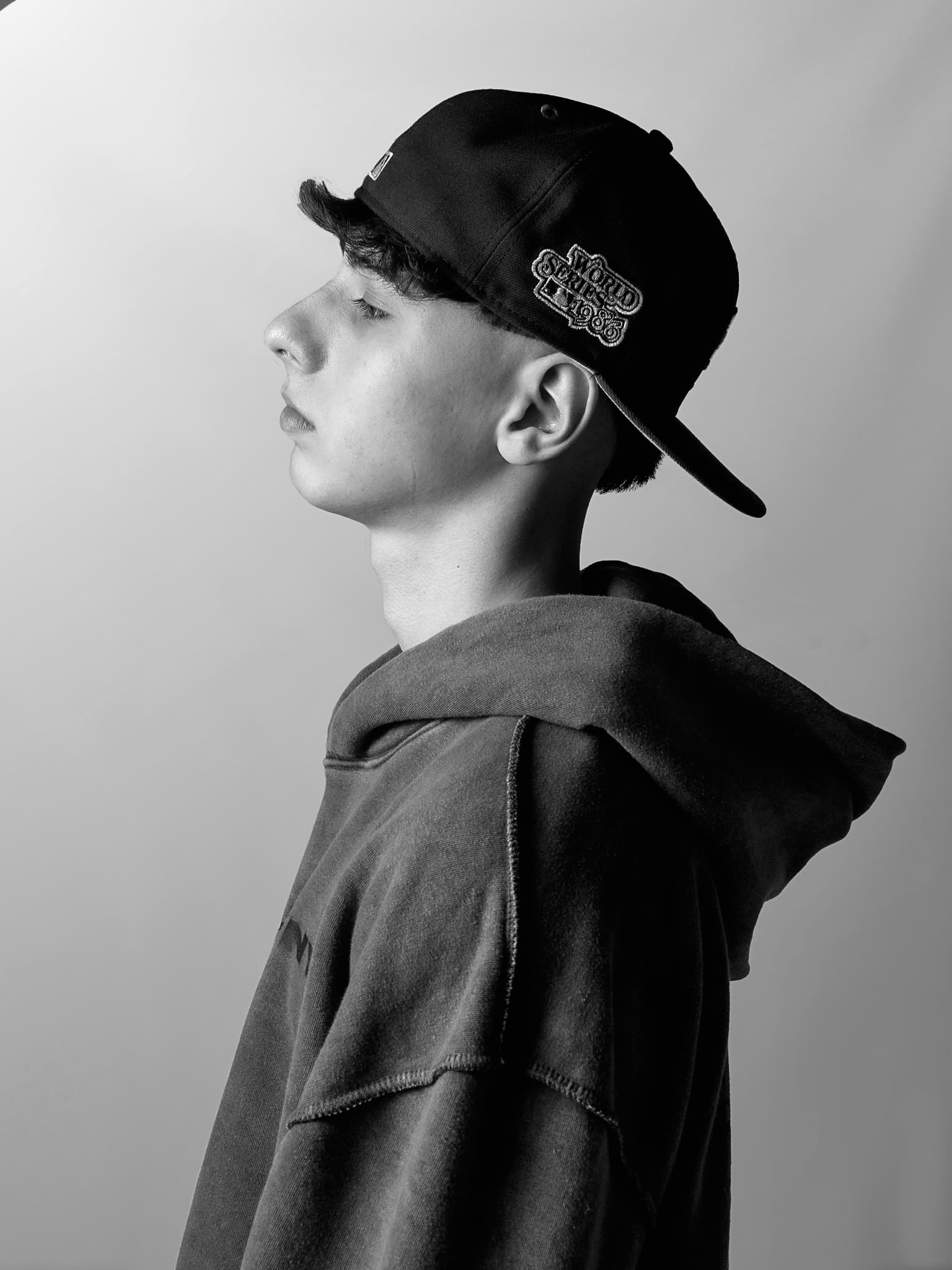 Retrato de perfil en blanco y negro, con gorra y buzo con capucha, sobre una pared clara
